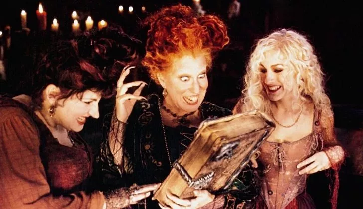 Сцена из фильма 'Фокус-покус' с тремя ведьмами, рассматривающими книгу заклинаний