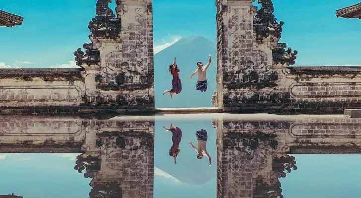 Два человека прыгают между двумя резными каменными столбами, а сцена отражается в отражении внизу